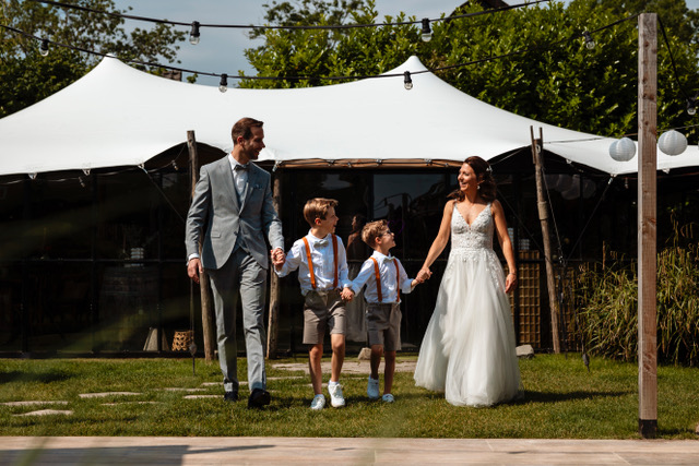 real bride sanneke – romantische A-lijn trouwjurk – bruid en bruidegom lopen samen met hun twee kinderen hand in hand over een zonnig grasveld bij een tentlocatie; vrolijke en liefdevolle familiesfeer